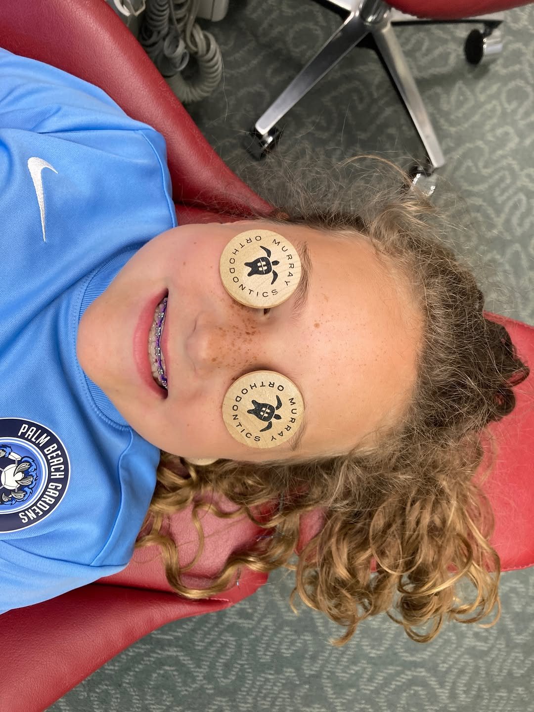 Smiling young braces patient in the treatment chair at Murray Orthodontics holding two turtle tokens over her eyes